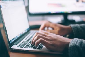 Hands typing at a Macbook Pro. A monitor is visible in the background.