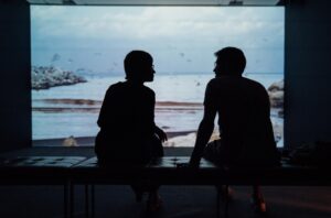 Photograph of two people talking facing a window and viewing the ocean on a cloudy day