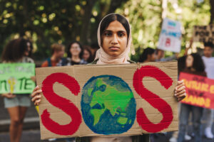 Photograph of a young person with a serious expression holding up a sign that reads “SOS” with a painting of Earth as the “O”