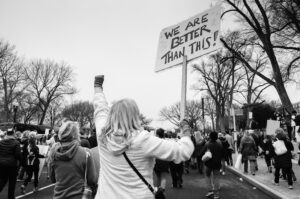 Black and white photograph of a climate march with a person holding a sign that reads “We are better than this!”