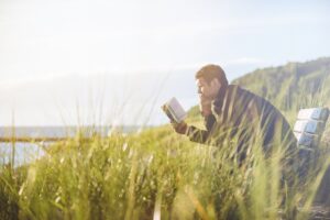 Photograph of a person reading on a bench surrounded by grass near a beach