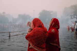 Two women in red sarees pray by a misty riverside during a crowded ritual.